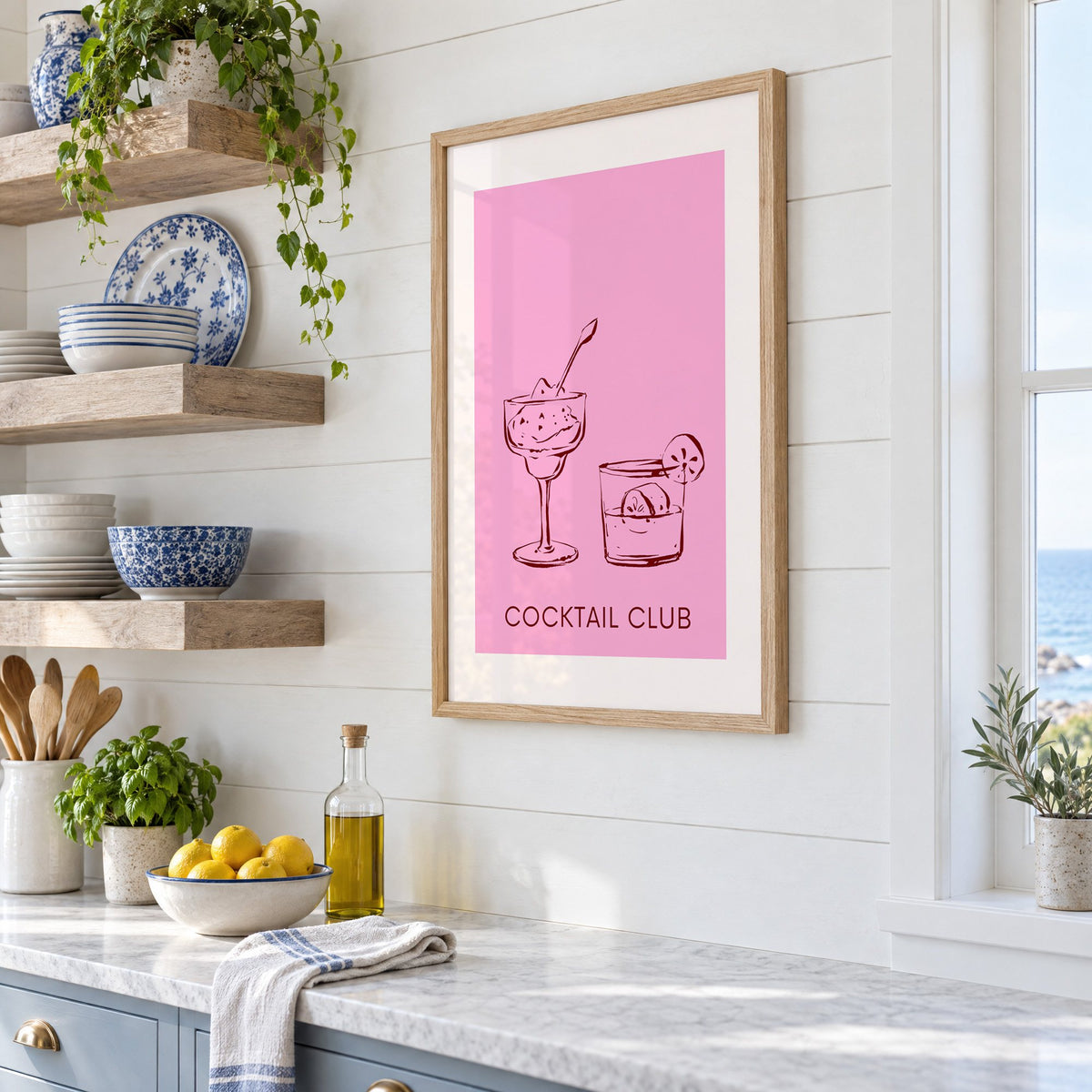 Cocktail Club sketched glasses pink print in an oak frame in a white coastal kitchen beside blue and white plates