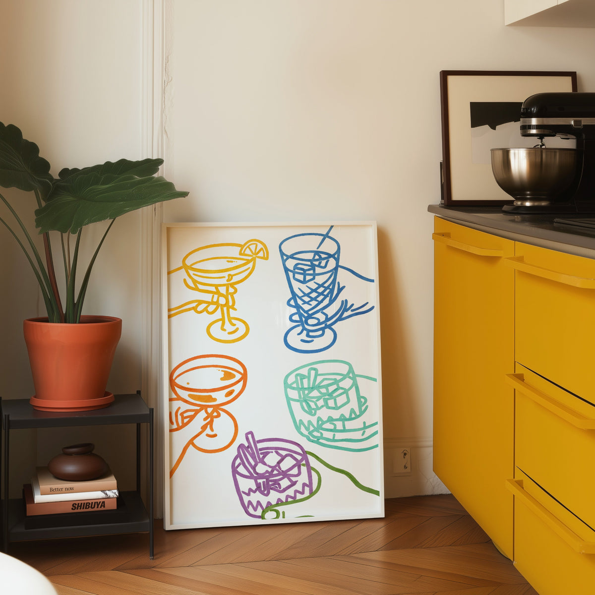 Cocktail glasses print in a white frame leaning on parquet beside a bright yellow kitchen