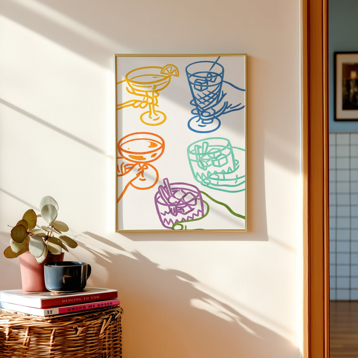 Cocktail glasses print in a gold frame above a basket of books and a pilea plant