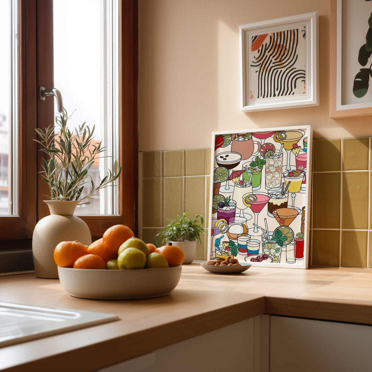 Dense cocktail glasses pattern print in a white frame leaning against olive green kitchen tiles beside a bowl of citrus
