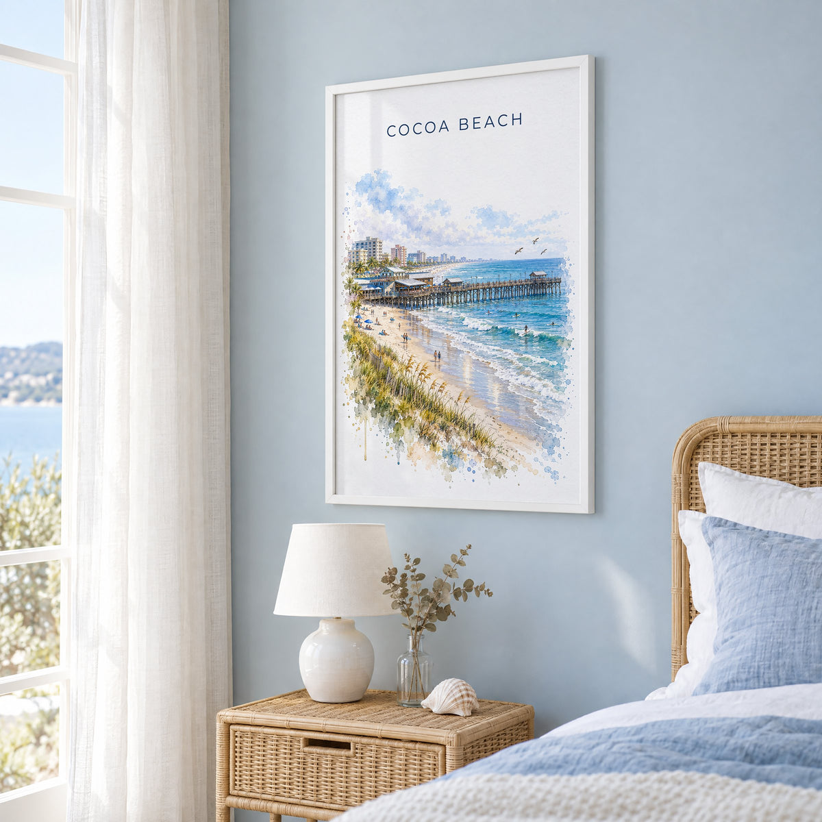 Cocoa Beach watercolor print in a white frame on a pale blue wall beside a rattan bed in a coastal bedroom