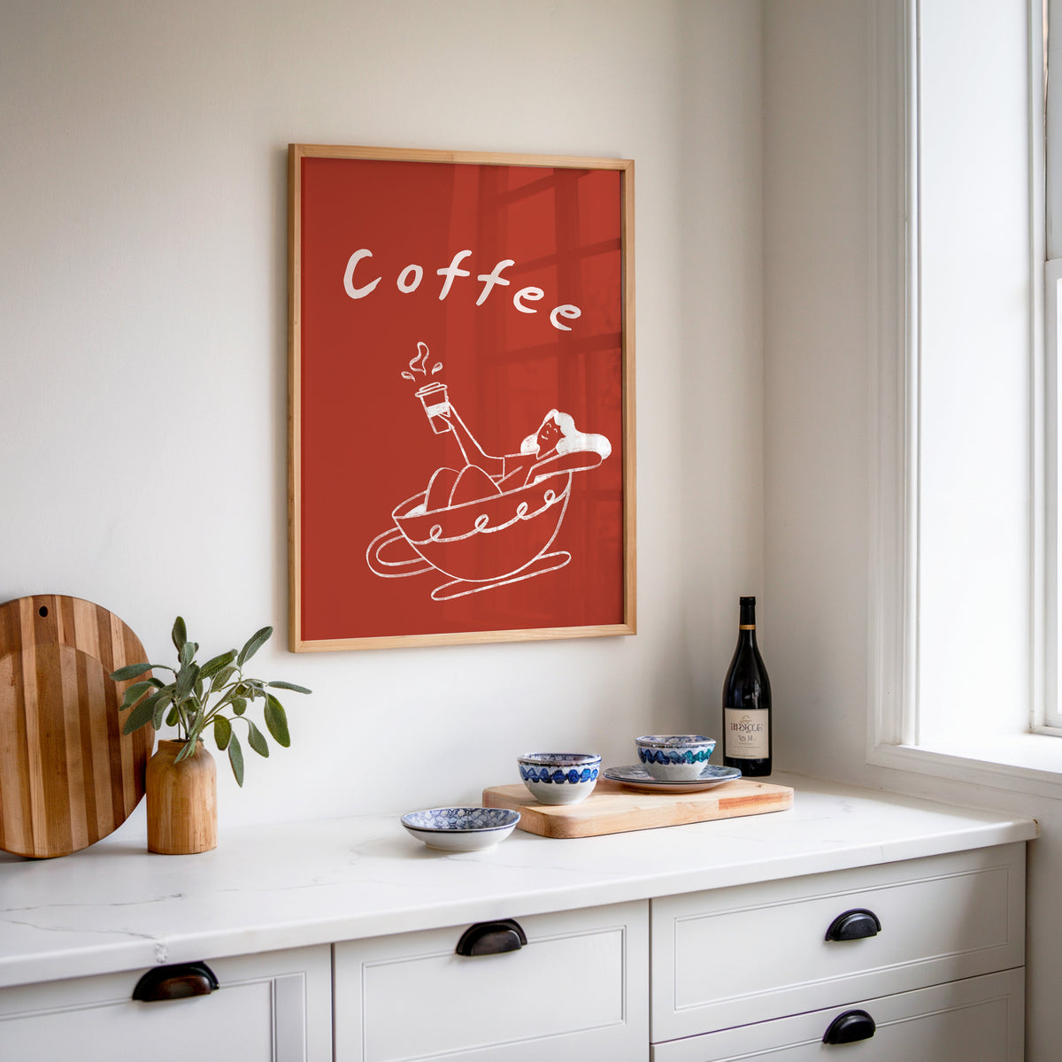 Coffee Bath print in an oak frame above a white sideboard with a bottle of wine