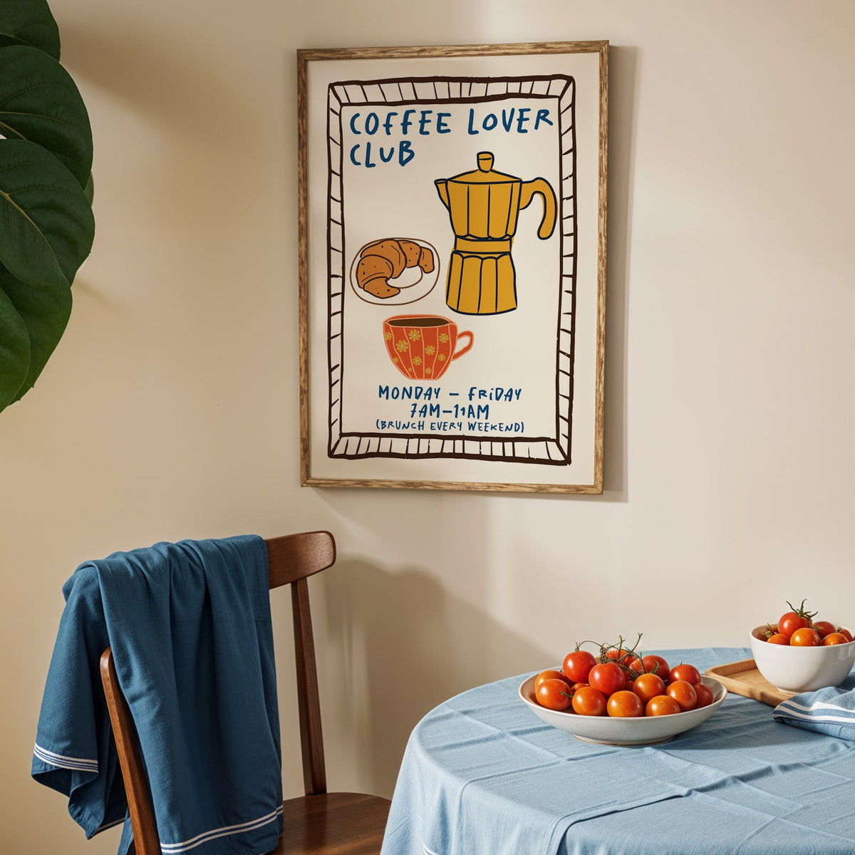 Coffee lover club moka pot print in an oak frame above a blue linen table with ripe tomatoes