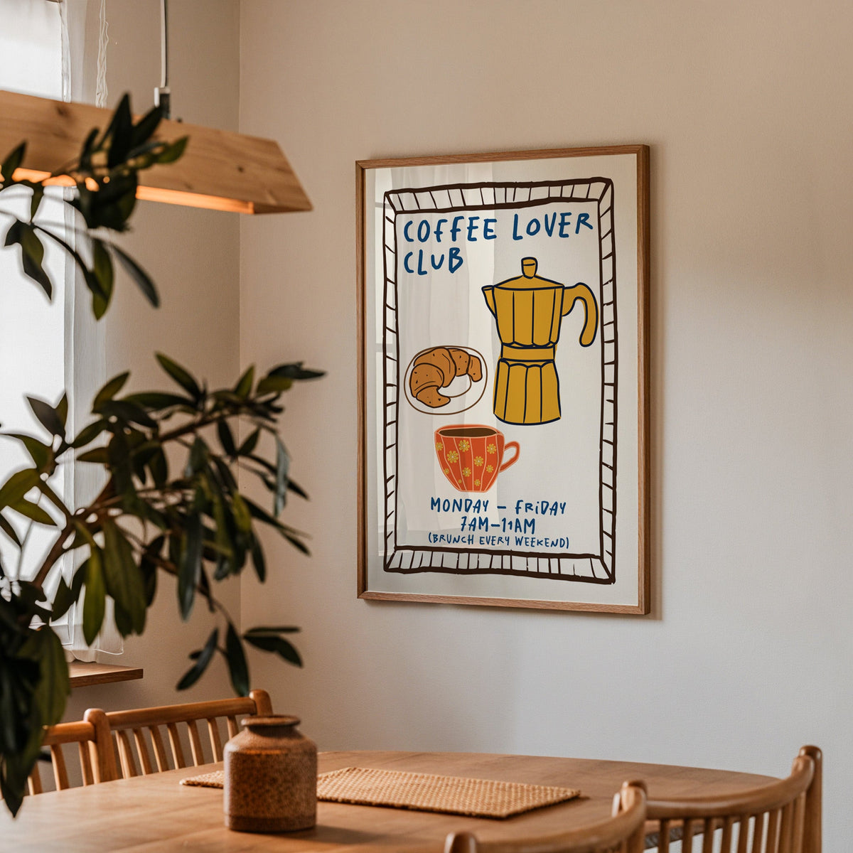 Coffee lover club moka pot print in a walnut frame above a dining table beside an olive tree