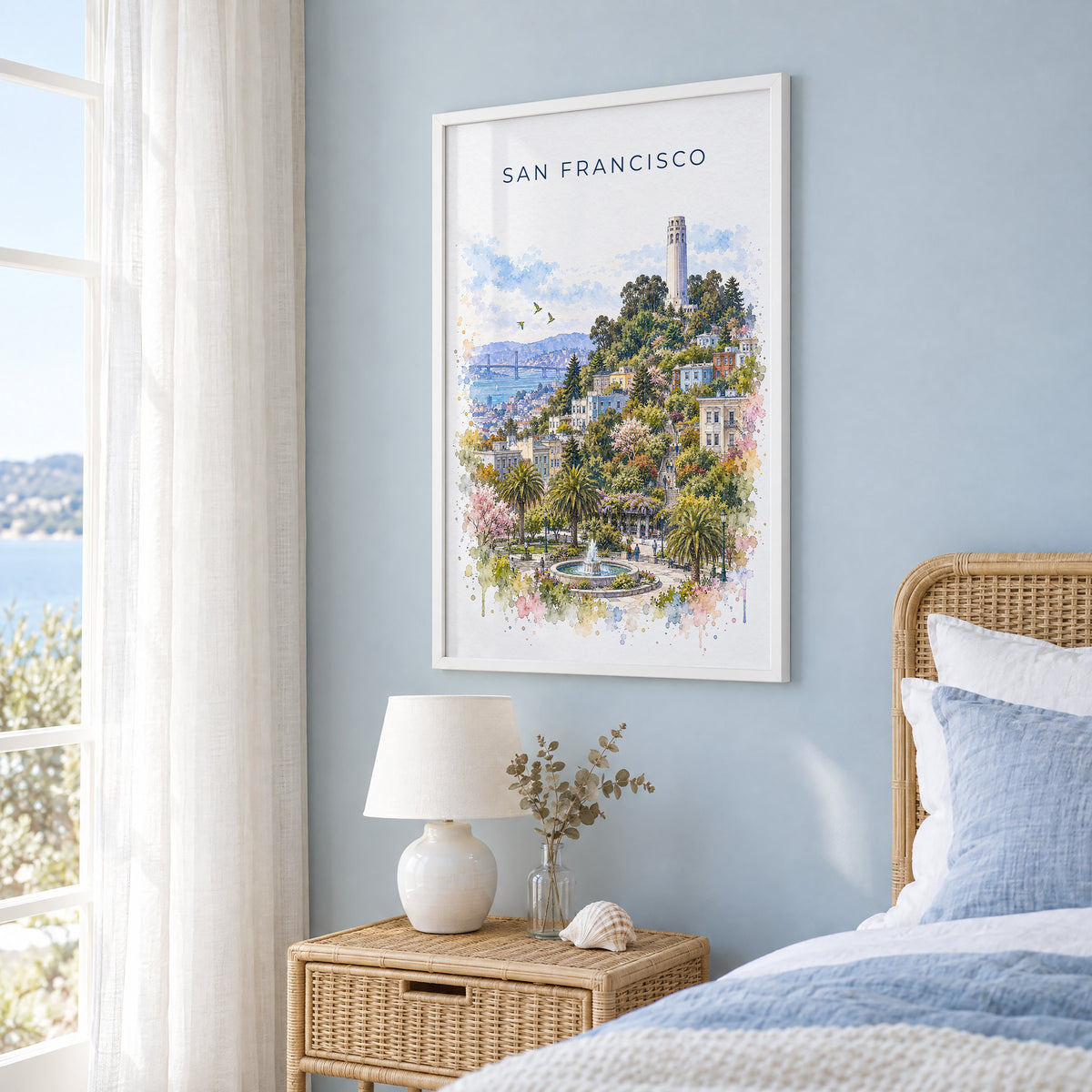 Coit Tower watercolor print in a white frame on a pale blue wall beside a rattan bed in a coastal bedroom