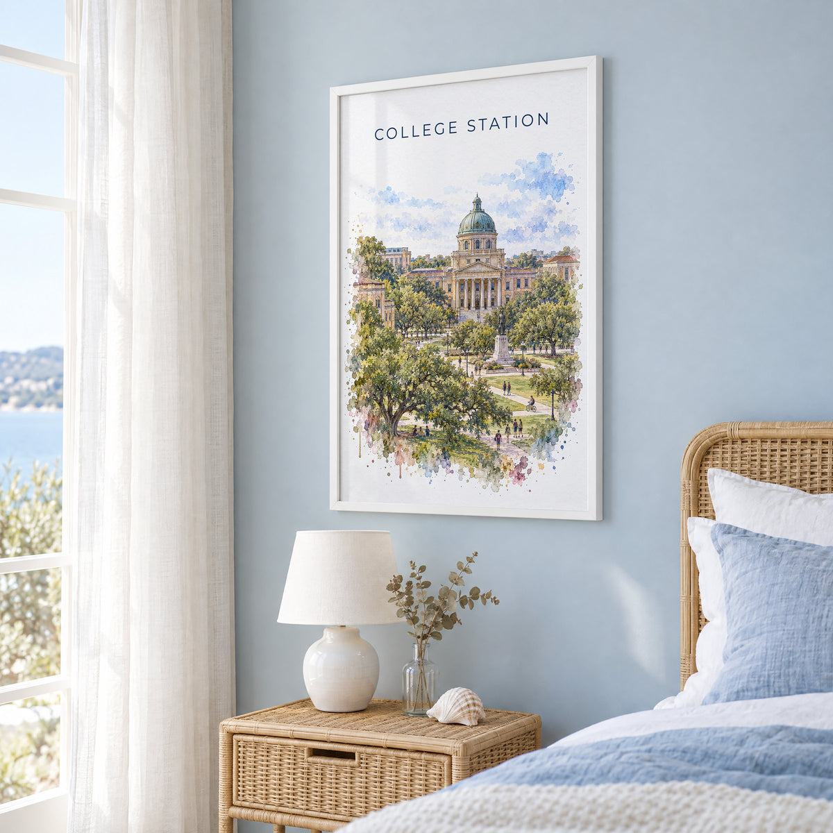 College Station watercolor print in a white frame on a pale blue wall beside a rattan bed in a coastal bedroom