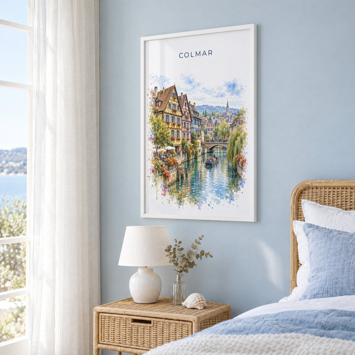 Colmar watercolor print in a white frame on a pale blue wall beside a rattan bed in a coastal bedroom