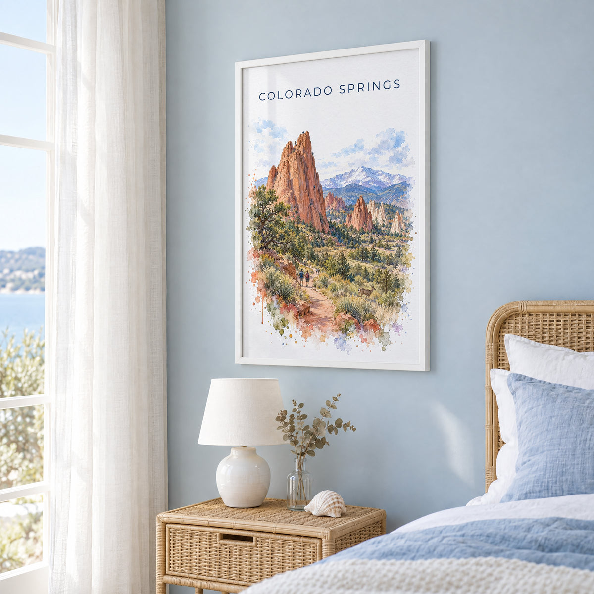 Colorado Springs watercolor print in a white frame on a pale blue wall beside a rattan bed in a coastal bedroom