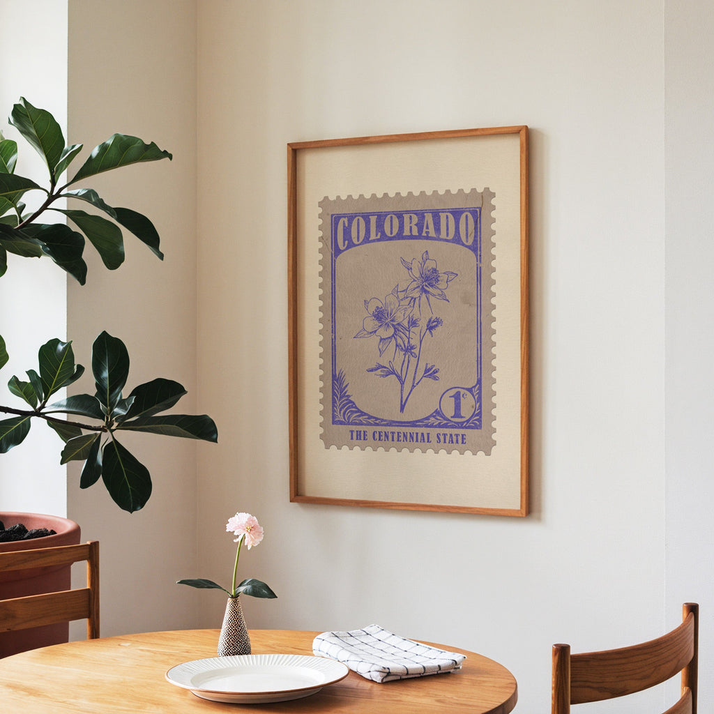 Colorado state flower print framed above a round wooden table with a single bloom in a vase and books