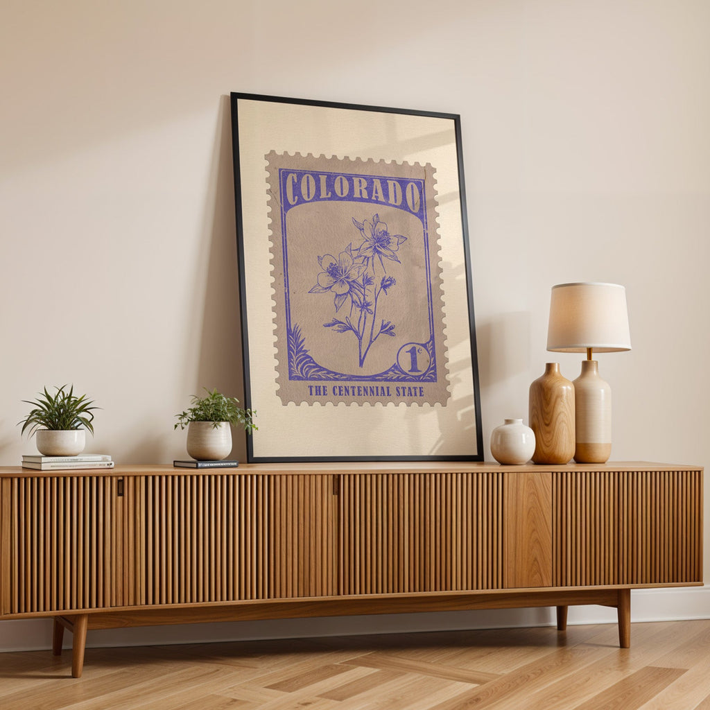 Colorado state flower print in a black frame leaning on a fluted wood credenza with vases and a lamp