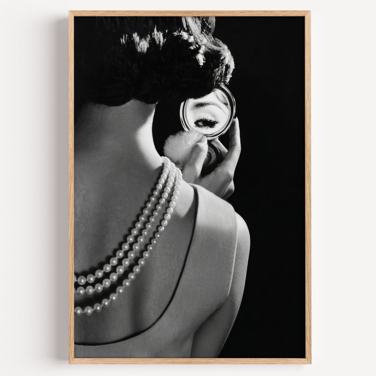 Woman from behind, eye in compact mirror, pearls print in a light oak frame on a plain white wall