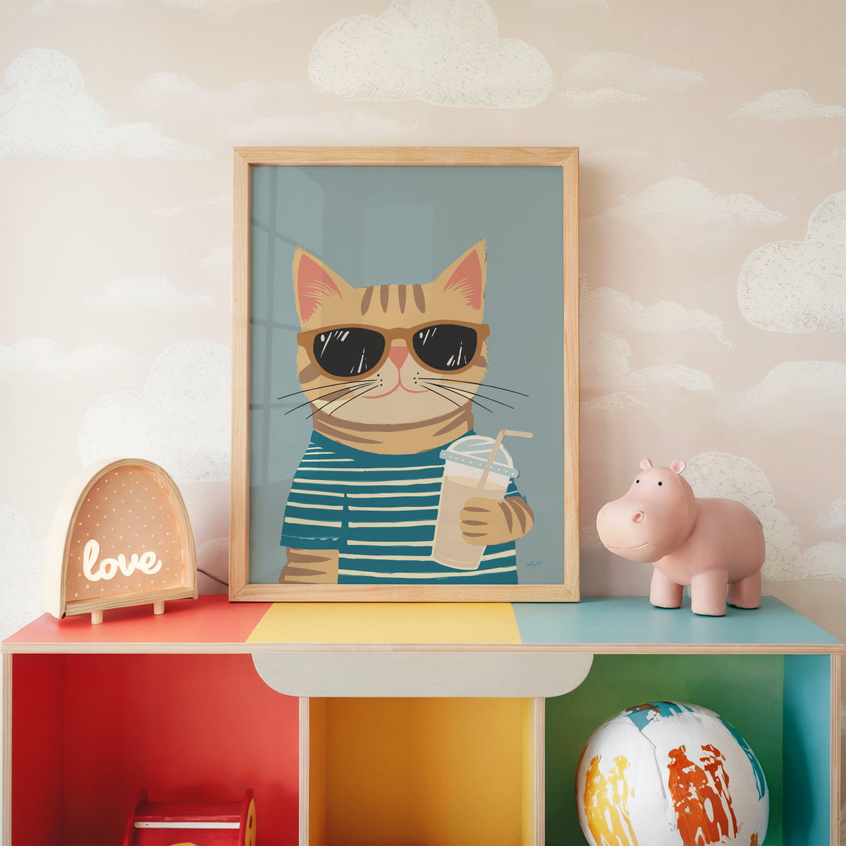 Orange tabby cat with iced drink nursery print in an oak frame on a rainbow toy shelf against cloud wallpaper