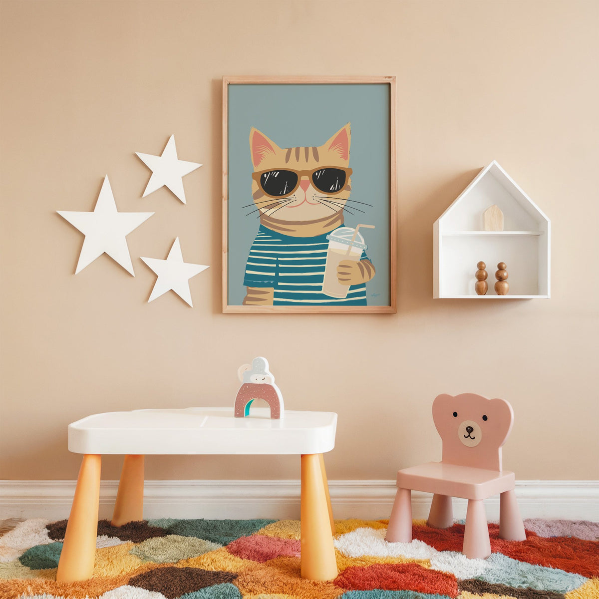 Orange tabby cat with iced drink nursery print in an oak frame on a peach wall above a kids table with wall stars
