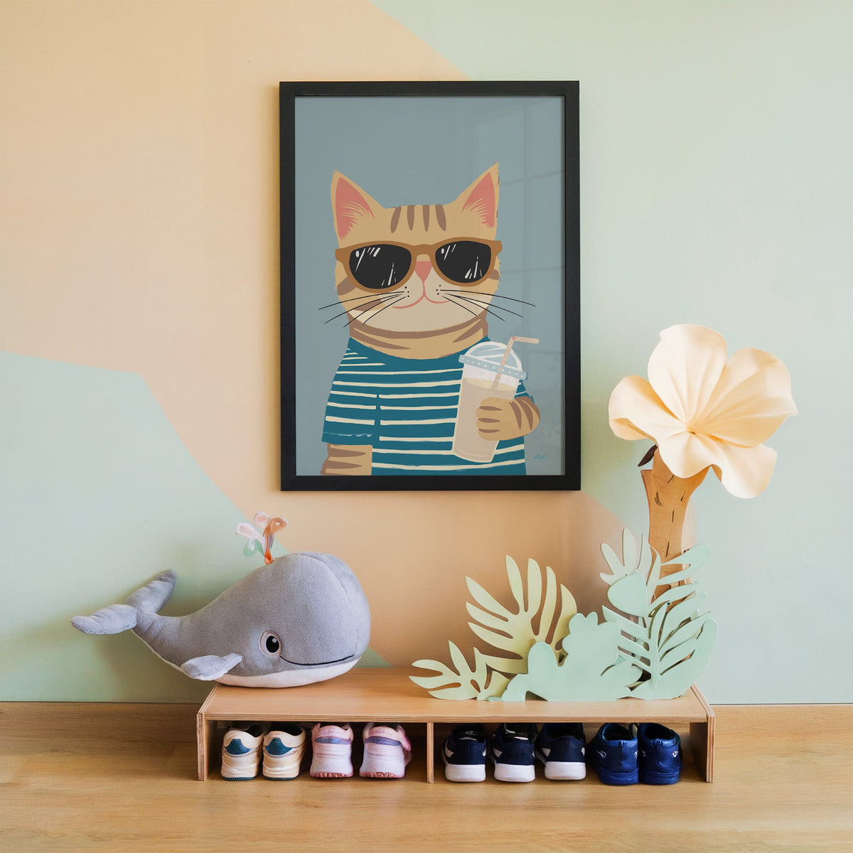 Orange tabby cat with iced drink nursery print in a black frame on a mint and peach wall above a whale plush