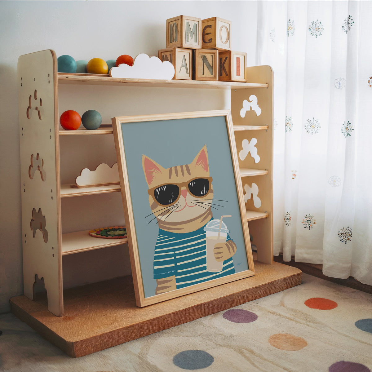 Orange tabby cat with iced drink nursery print in an oak frame leaning on a toy shelf with wooden letter blocks