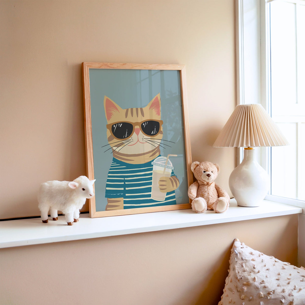 Orange tabby cat with iced drink nursery print in an oak frame on a window ledge with a toy lamb and teddy