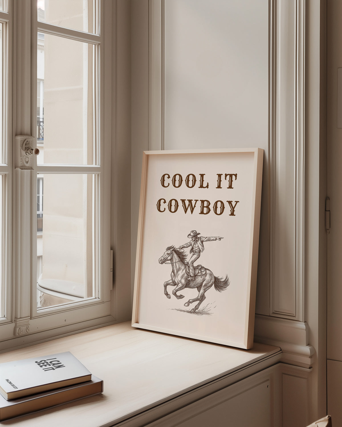 Cool It Cowboy print in a pale wood frame on a sunlit window ledge
