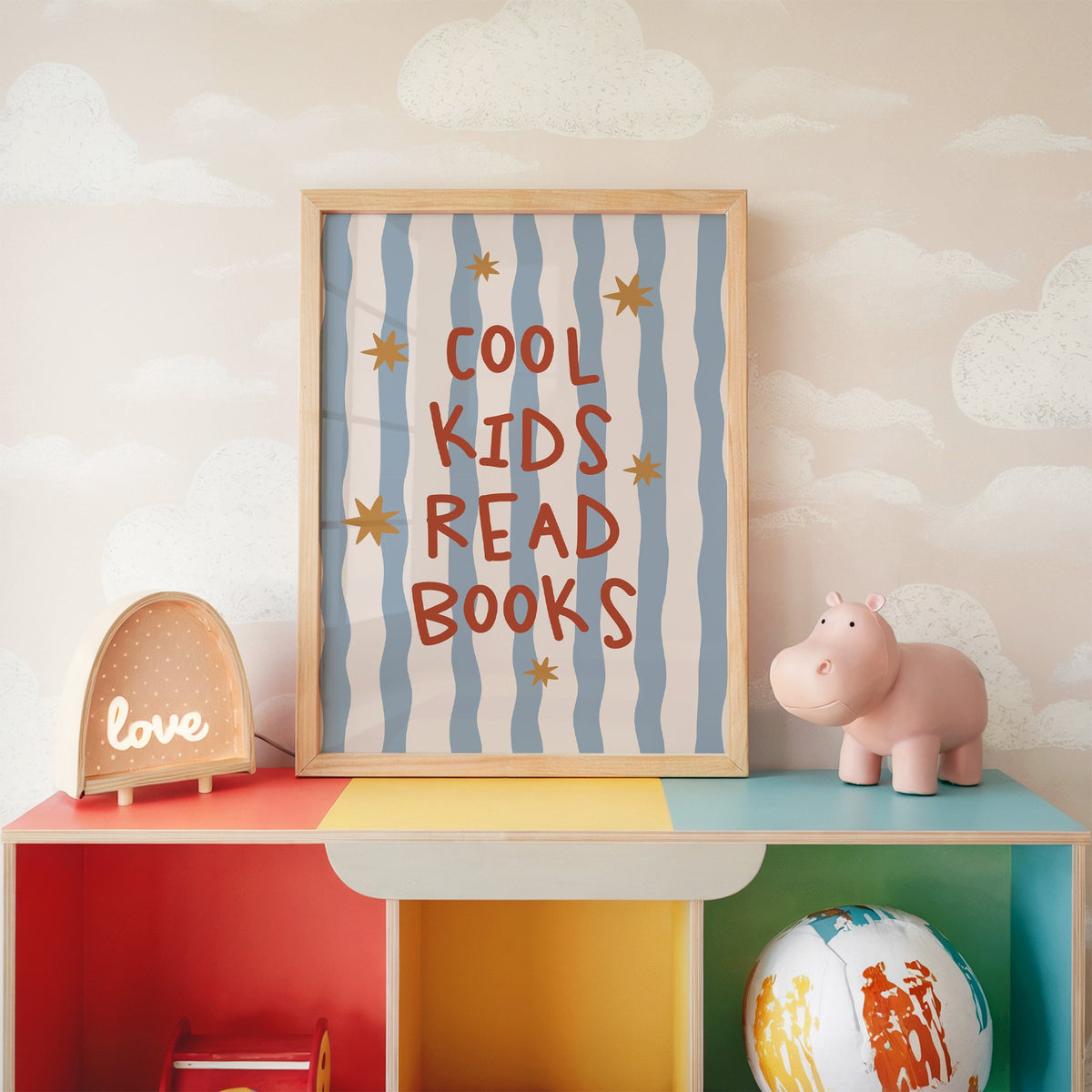 Cool kids read books gold stars nursery print in an oak frame on a rainbow toy shelf against cloud wallpaper