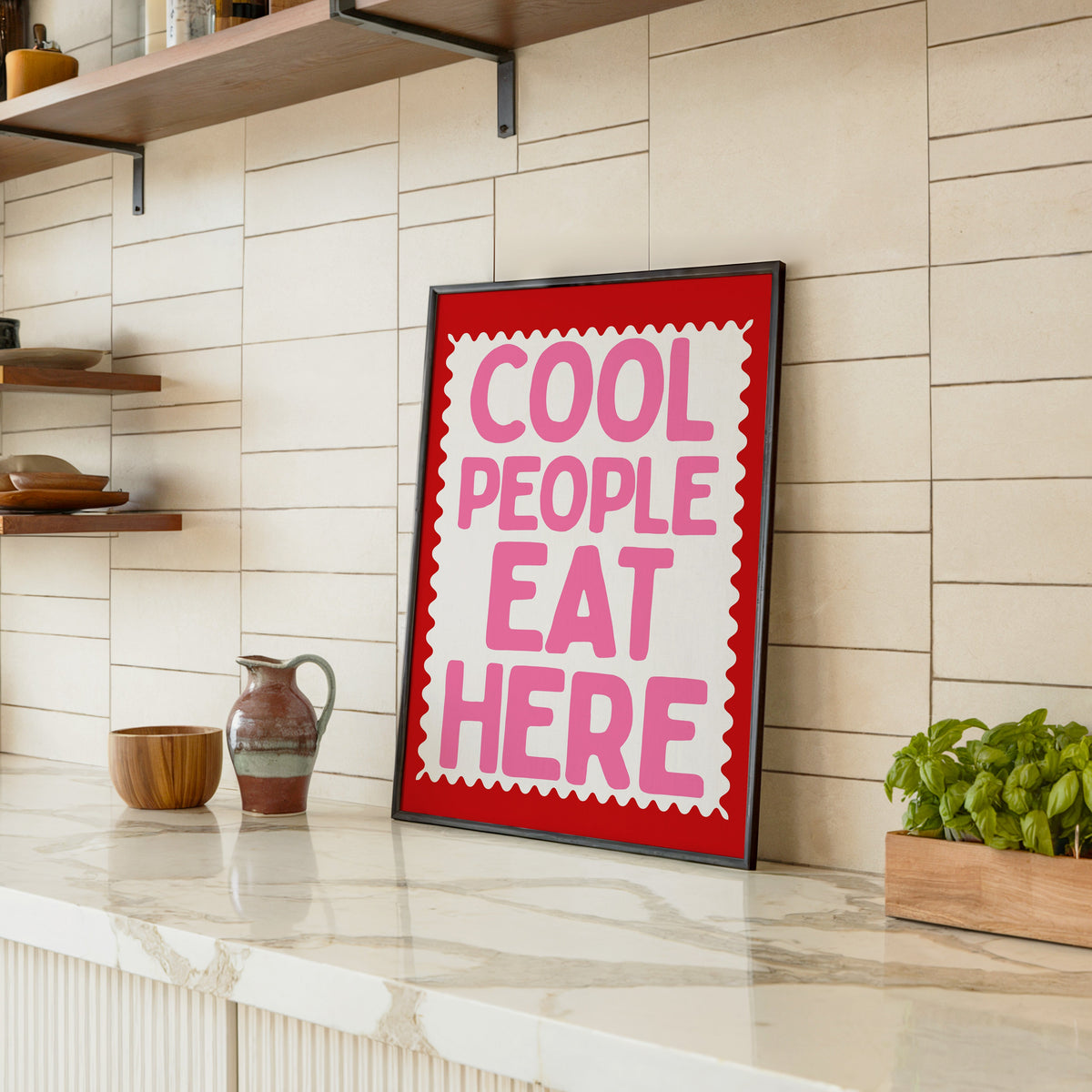 Cool People Eat Here print in a black frame leaning on a marble counter in a tiled kitchen