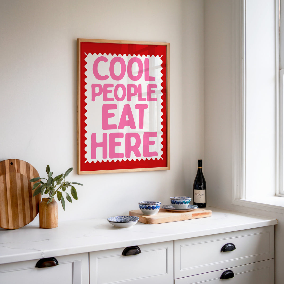 Cool People Eat Here print in an oak frame above a white sideboard with a bottle of wine