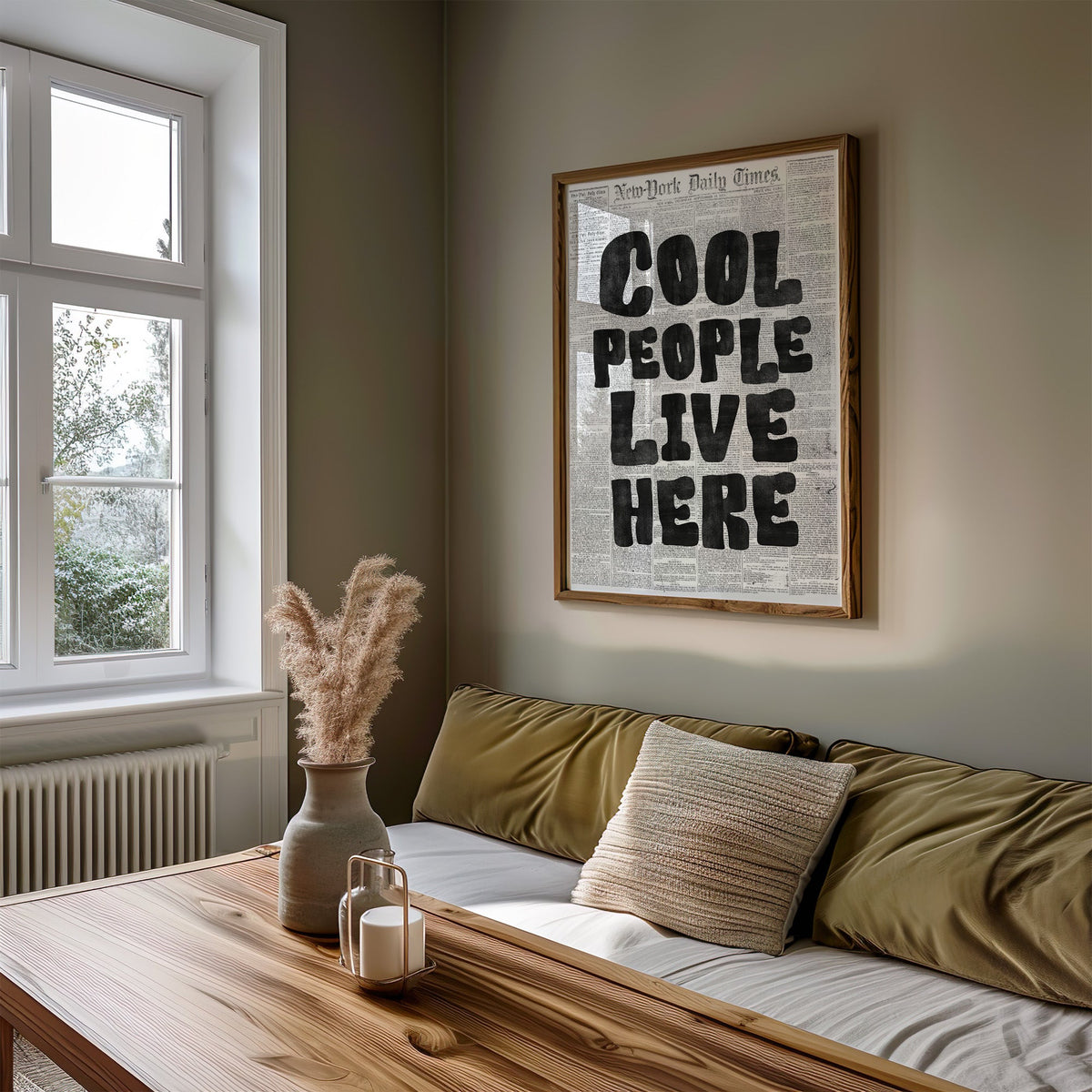 Cool People Live Here lettering on old newspaper print in a walnut frame above an olive daybed by a window
