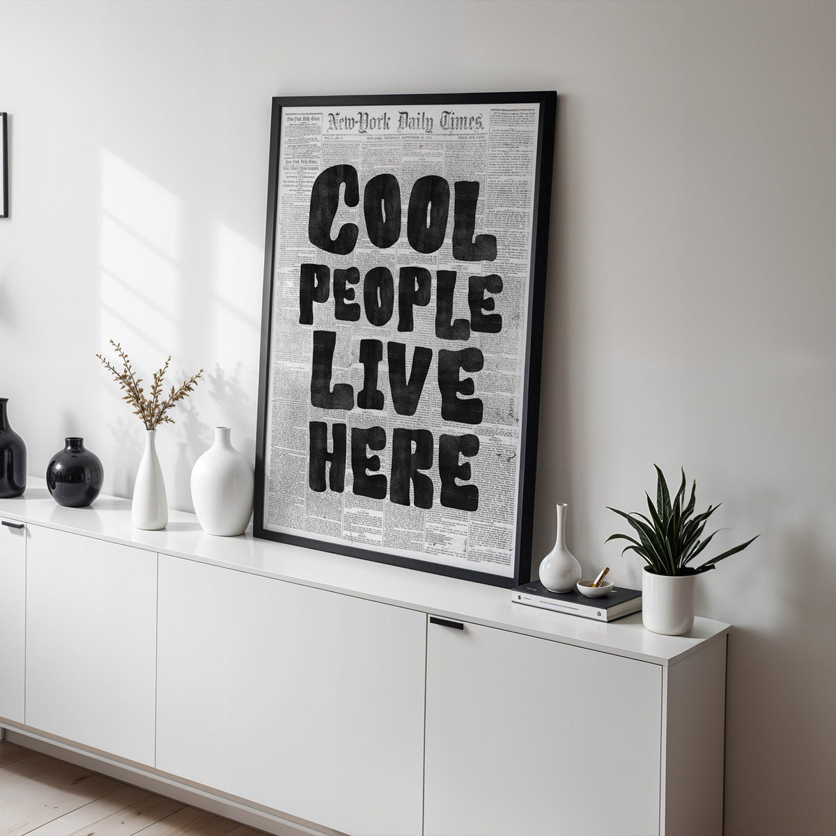 Cool People Live Here lettering on old newspaper print in a black frame on a white sideboard with black and white vases