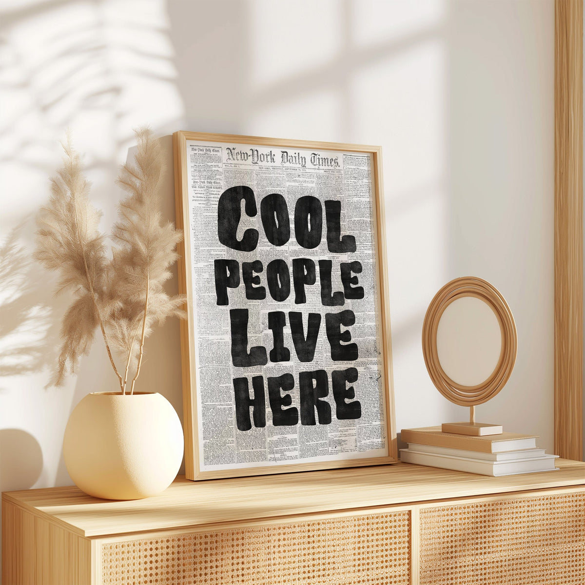 Cool People Live Here lettering on old newspaper print in an oak frame on a rattan sideboard beside pampas grass