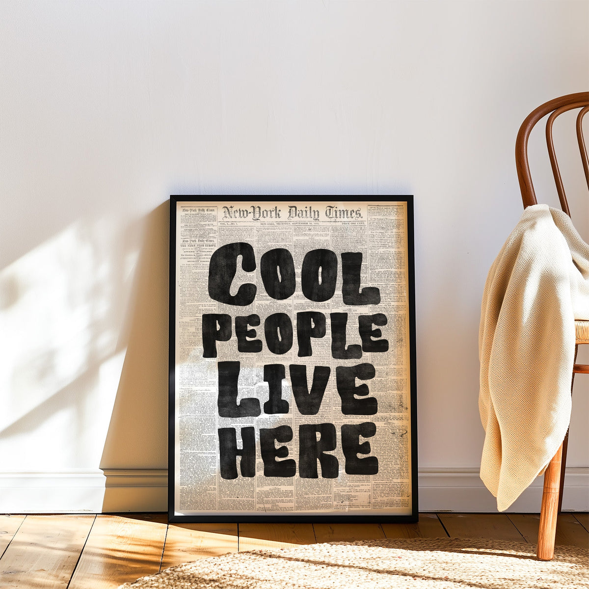 Cool People Live Here lettering on old newspaper print in a black frame leaning on a wood floor beside a bentwood chair