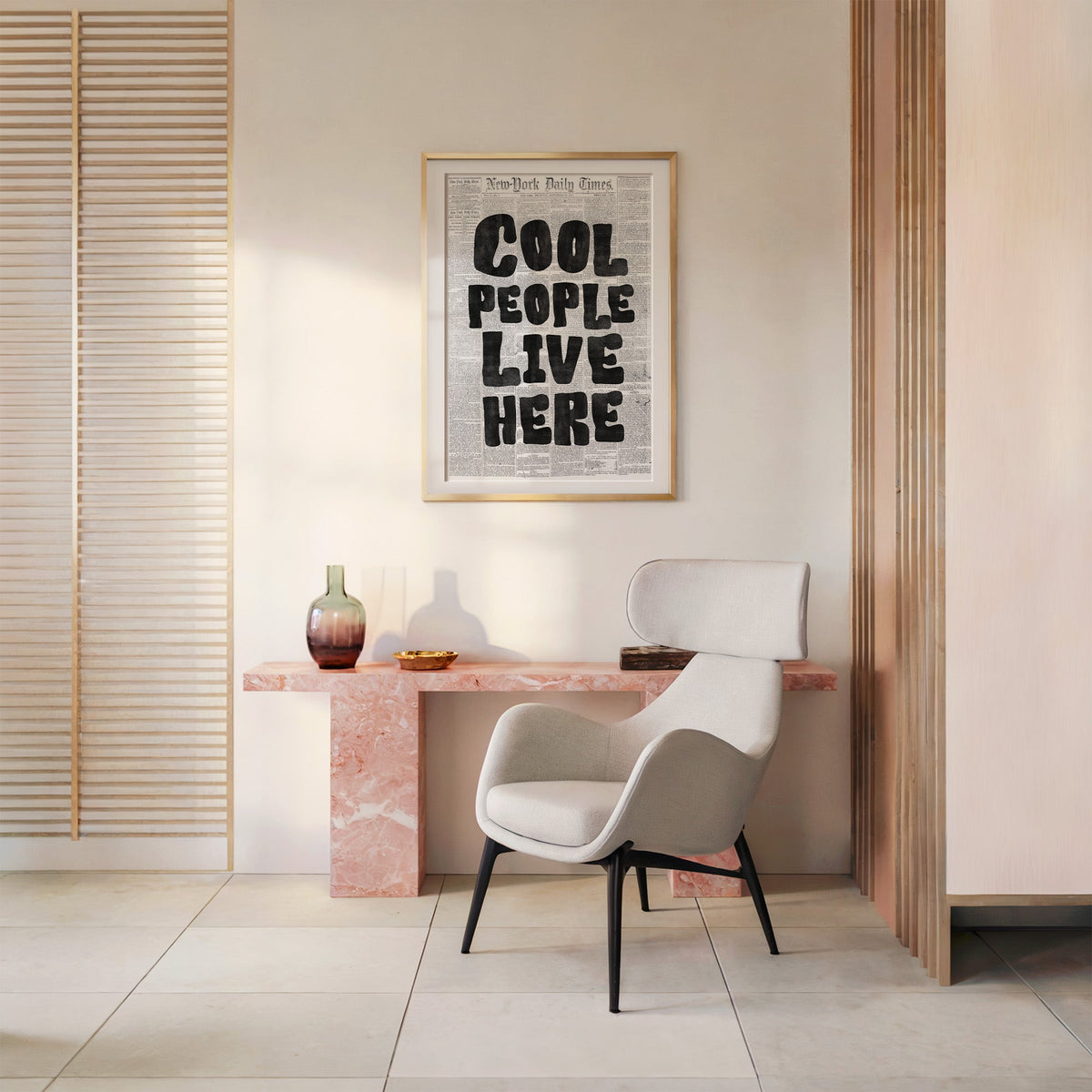 Cool People Live Here lettering on old newspaper print shown in a gold frame above a pink marble console and armchair