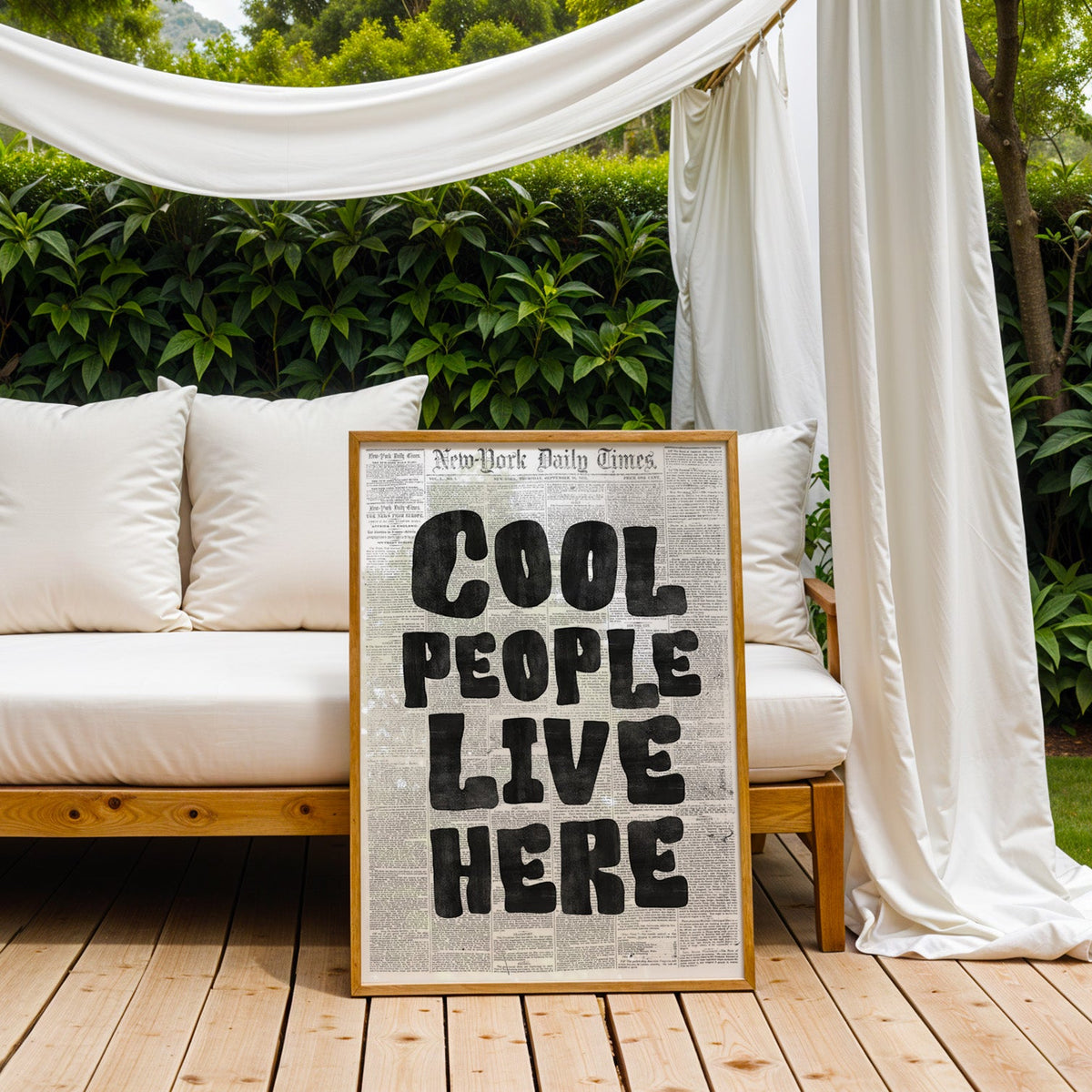 Cool People Live Here lettering on old newspaper print in an oak frame on an outdoor daybed under white drapes
