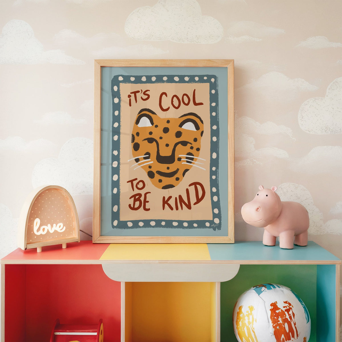Cool to be kind cat quote nursery print in an oak frame on a rainbow toy shelf against cloud wallpaper