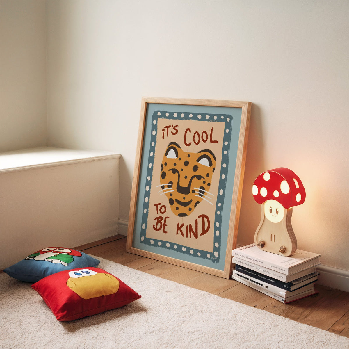 Cool to be kind cat quote nursery print in an oak frame leaning on a rug beside a mushroom lamp and floor cushions