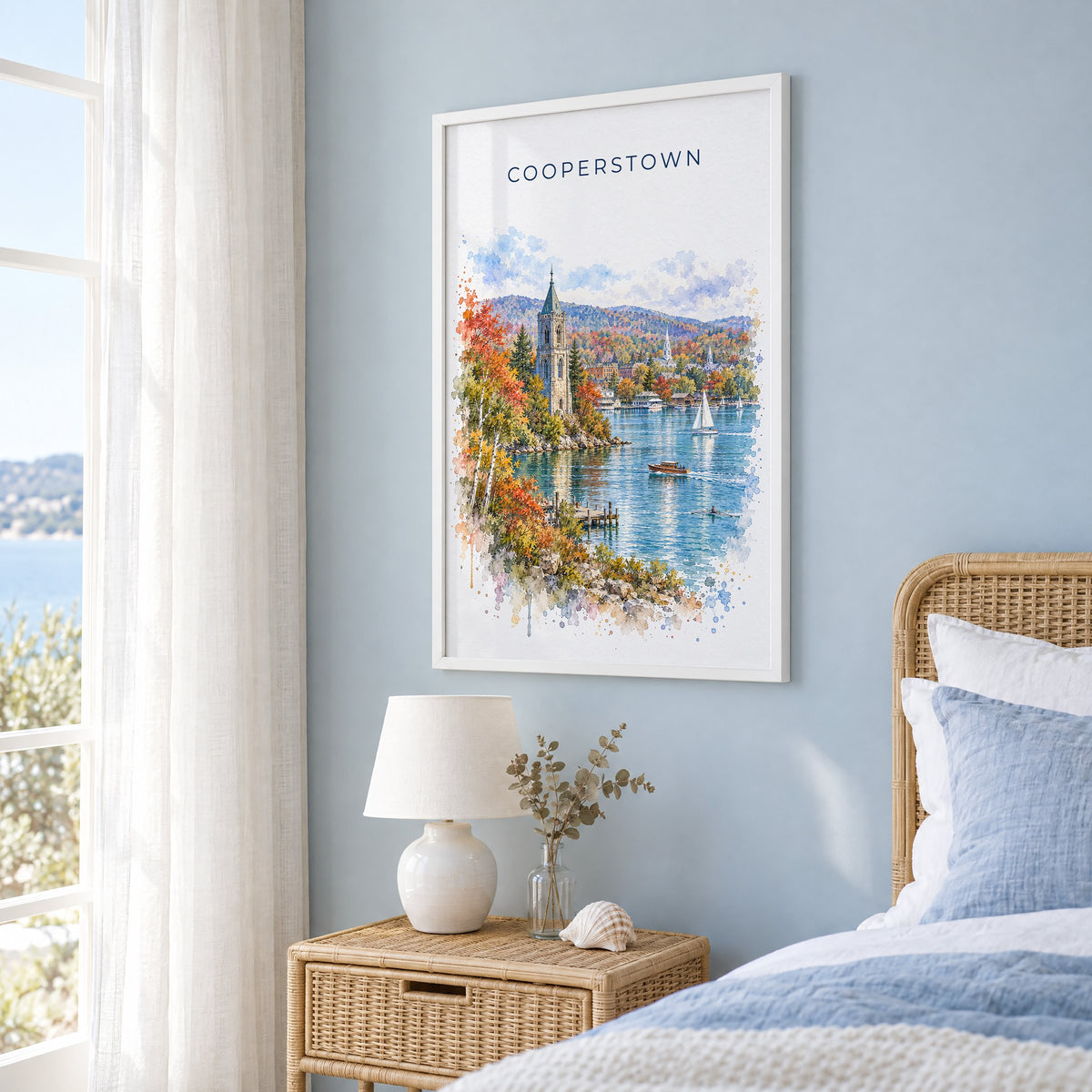 Cooperstown watercolor print in a white frame on a pale blue wall beside a rattan bed in a coastal bedroom
