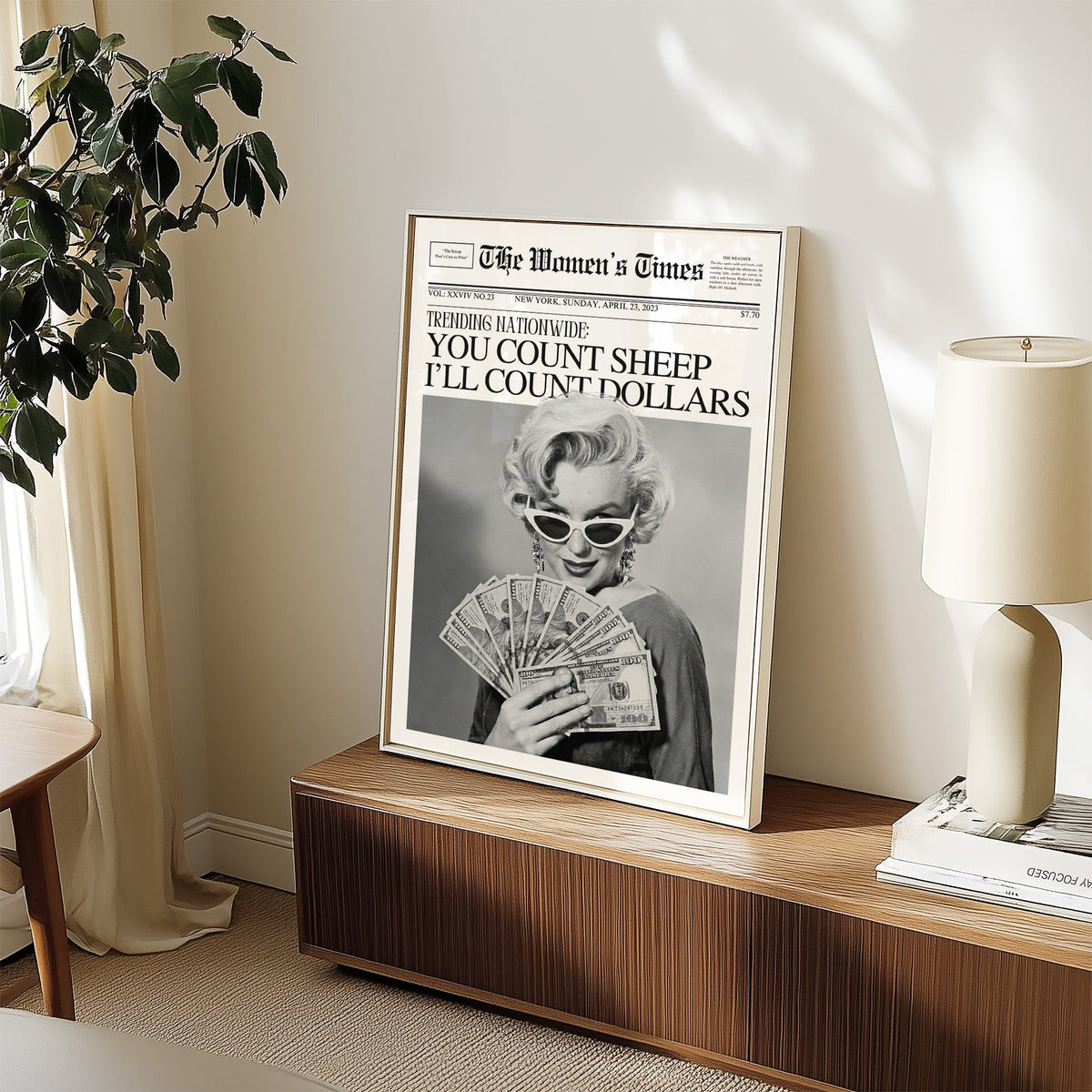 Woman fanning dollar bills mock front page print in a white frame on a walnut sideboard beside a ceramic lamp