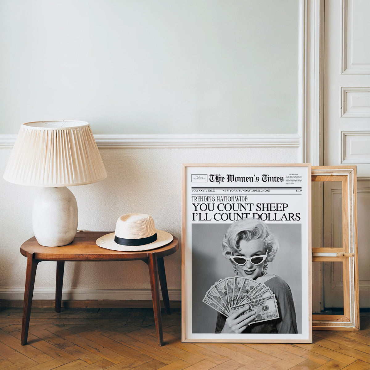 Woman fanning dollar bills mock front page print in a pine frame on a wood floor beside a lamp and straw hat