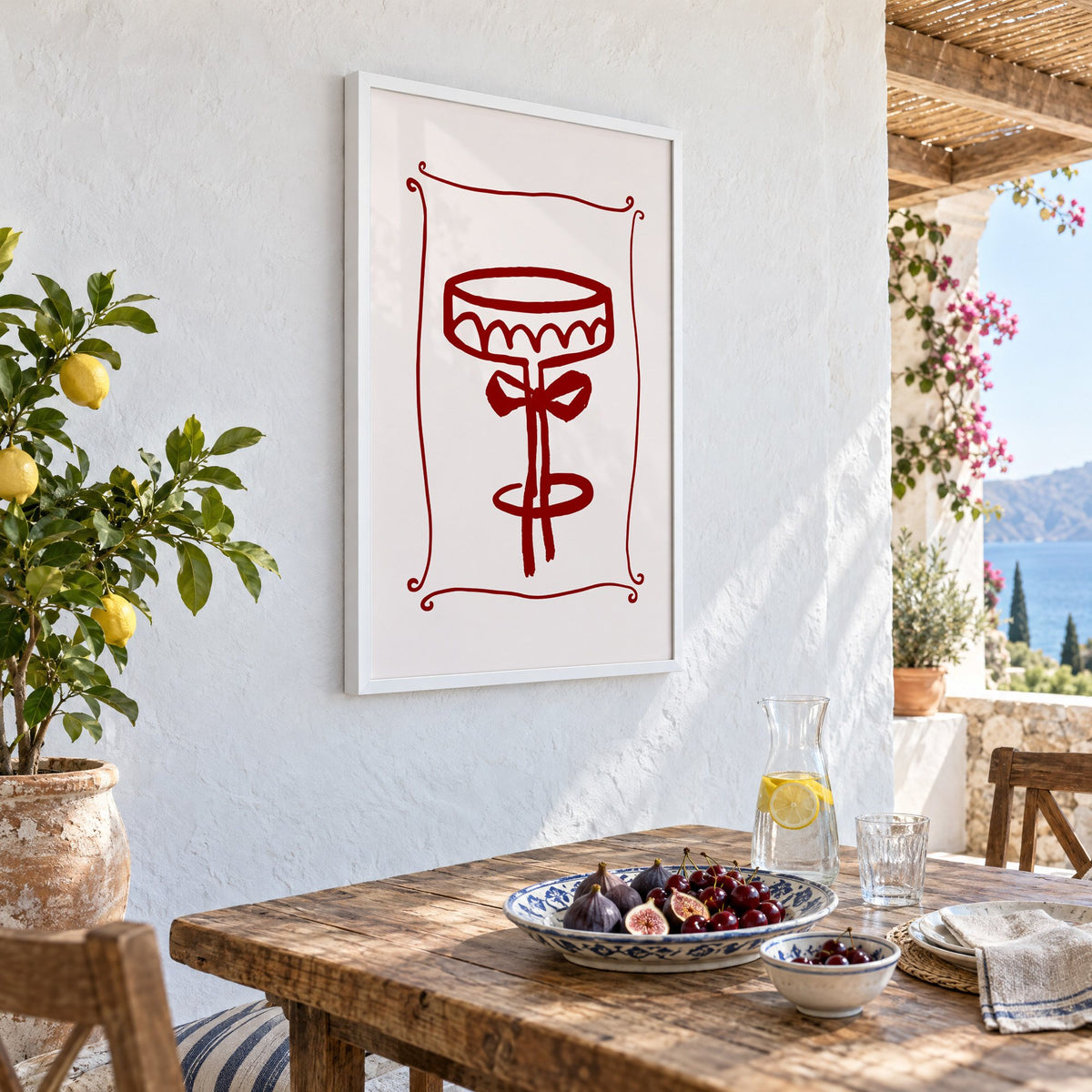 burgundy brush coupe glass with bow print in a white frame on a whitewashed terrace wall above a lunch table with figs and cherries, seen at an angle