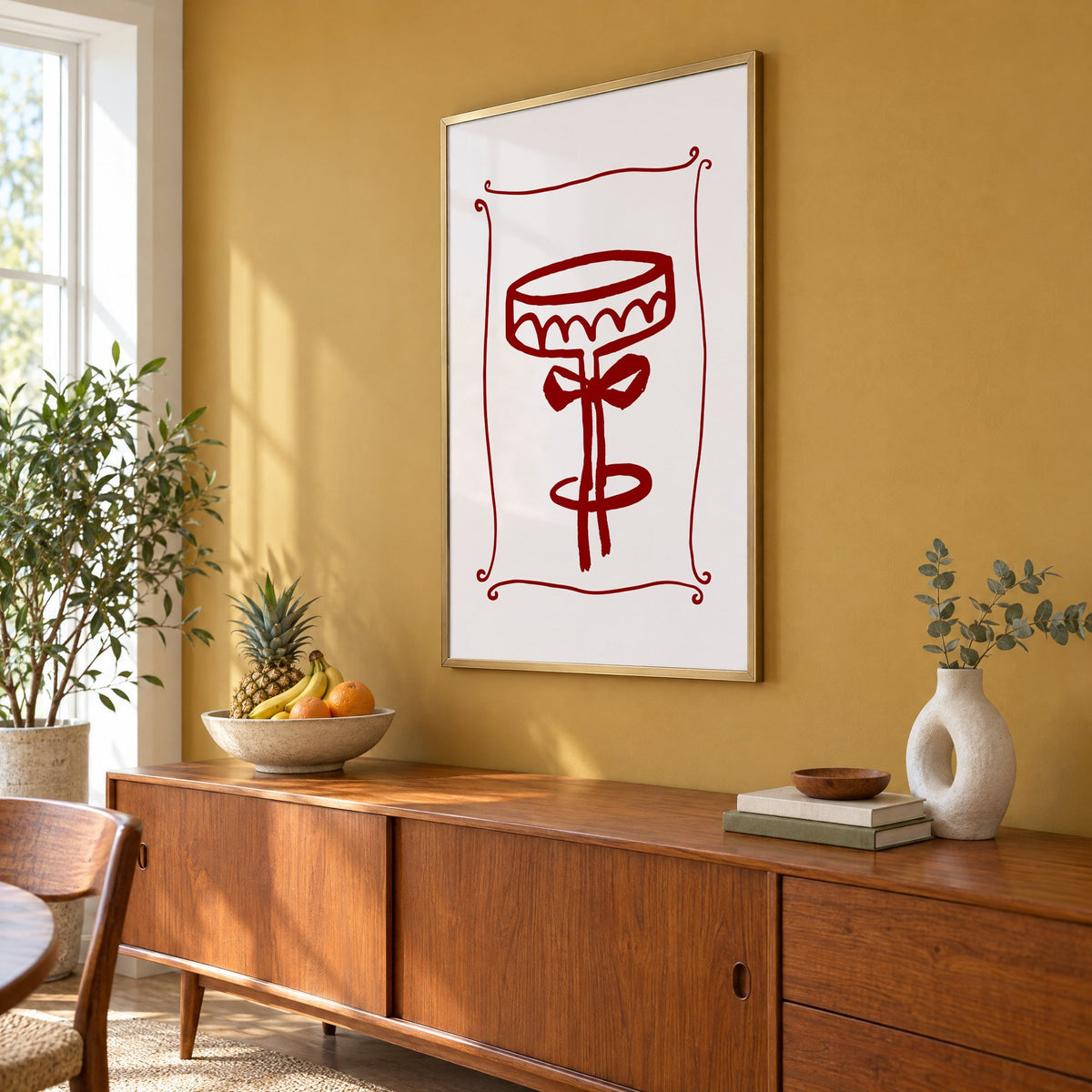 burgundy brush coupe glass with bow print in a brass frame on a mustard wall above a teak sideboard with a fruit bowl, seen at an angle