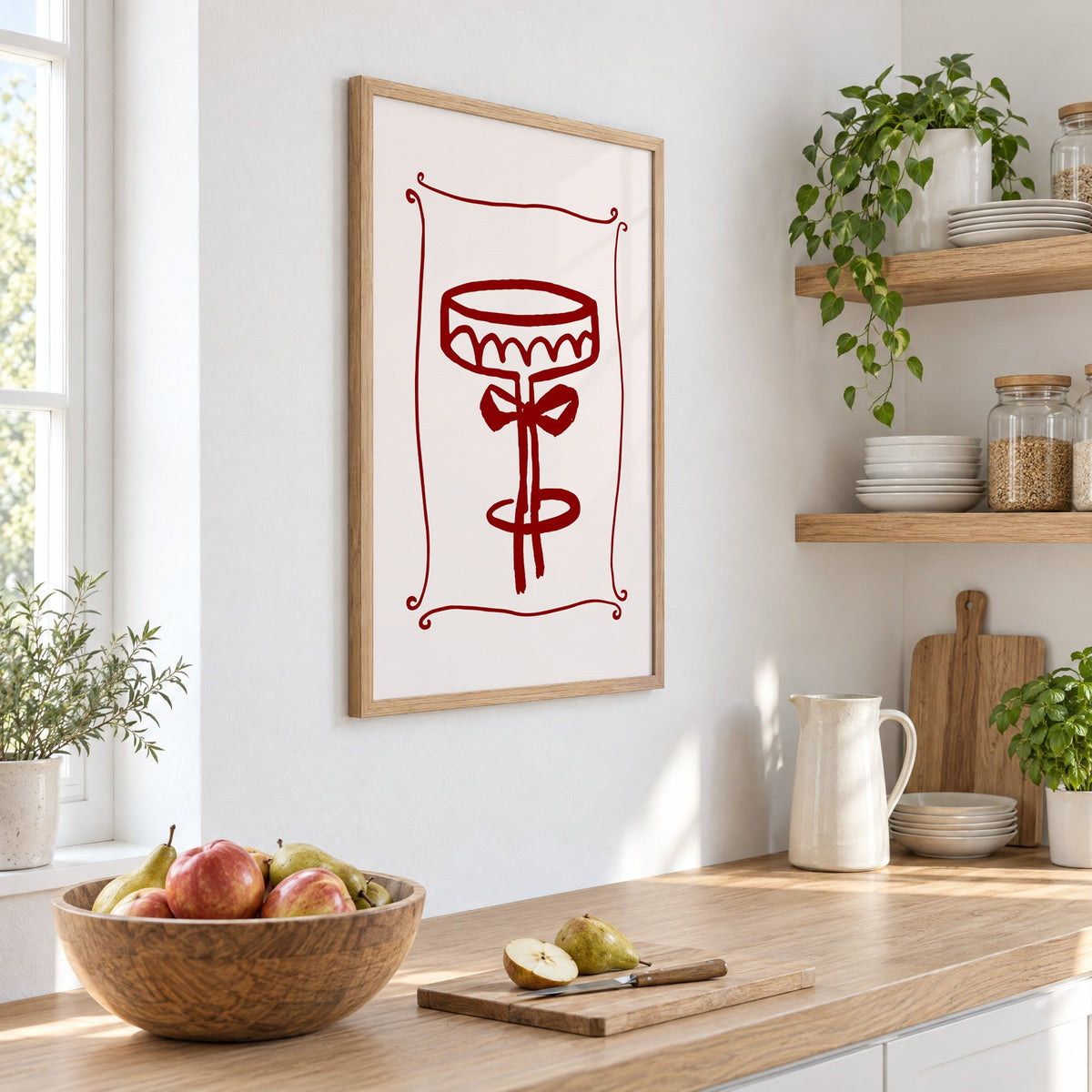 burgundy brush coupe glass with bow print in an oak frame on a white kitchen wall above a counter with apples and pears, seen at an angle