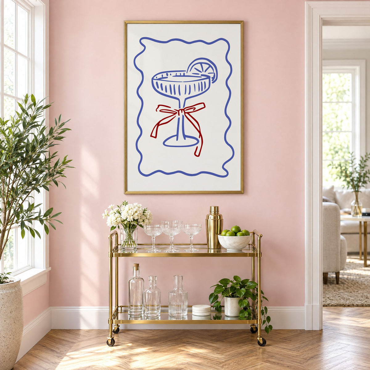 Blue coupe glass with red bow print in a brass frame on a blush pink wall above a gold bar cart with glassware