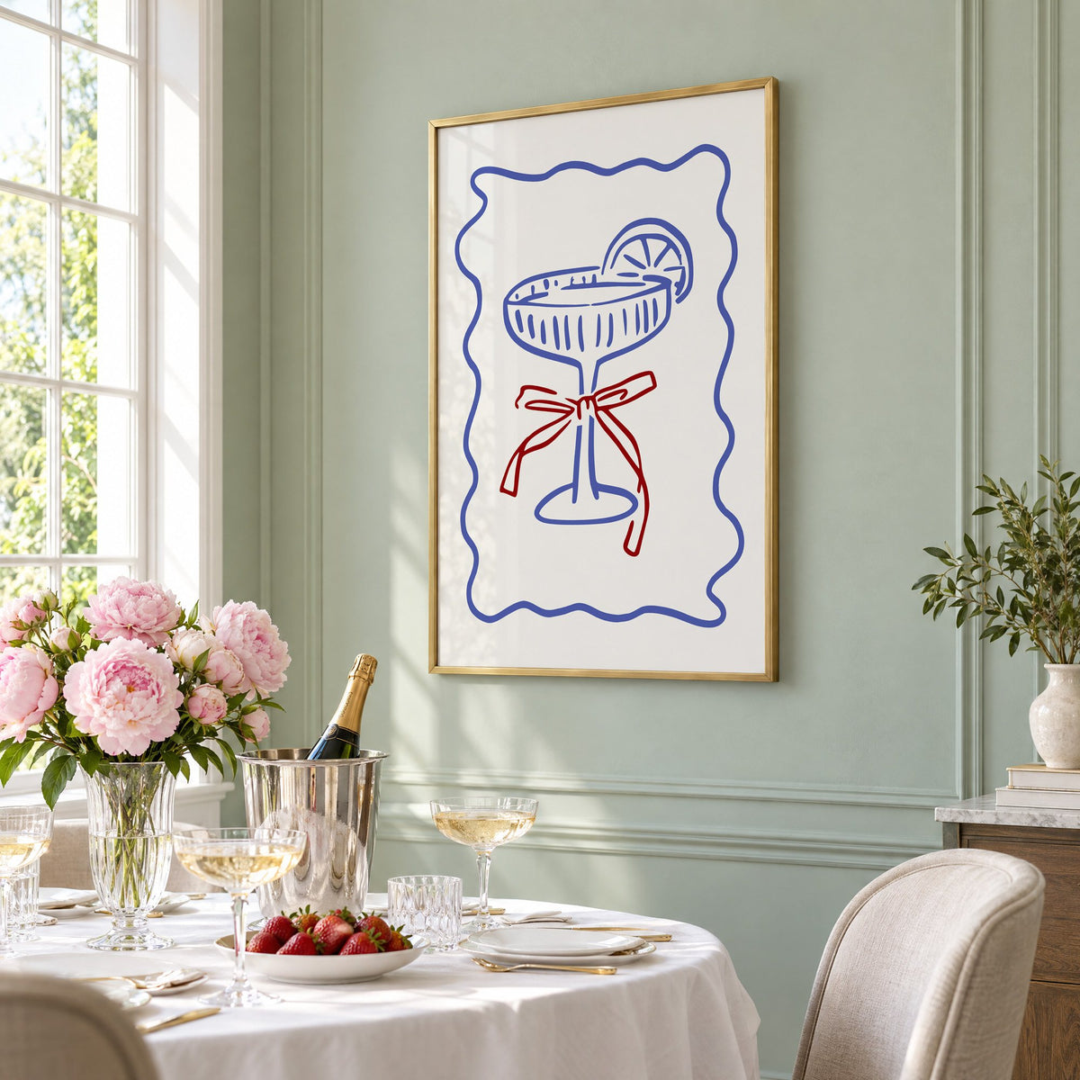 Blue coupe glass with red bow print in a brass frame on a sage wall above a brunch table with champagne and peonies, seen at an angle