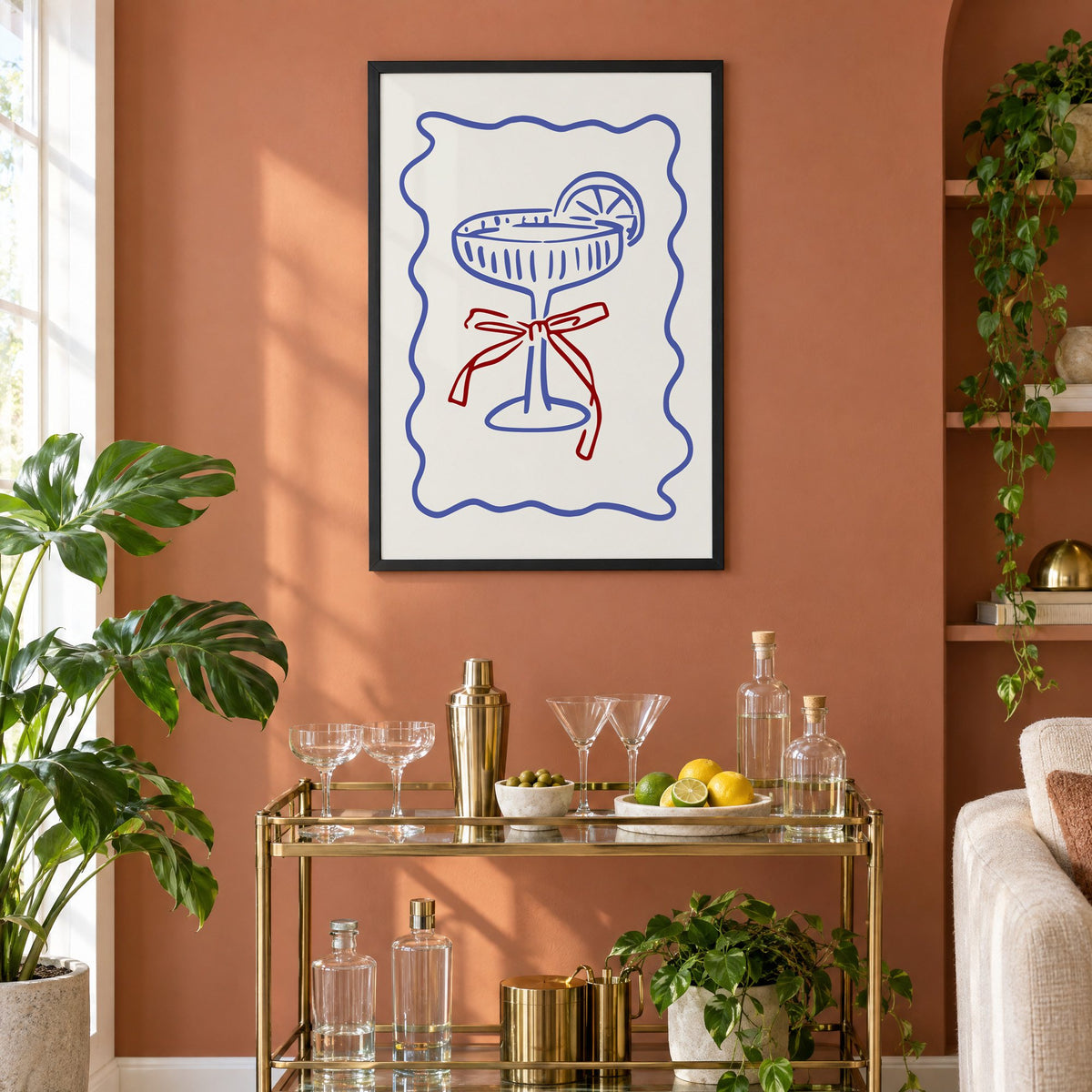 Blue coupe glass with red bow print in a black frame on a terracotta wall above a brass bar cart with coupes and citrus