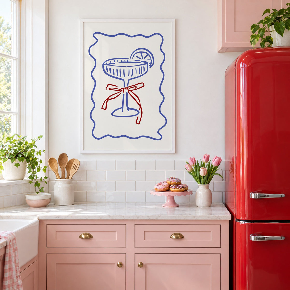 Blue coupe glass with red bow print in a white frame above pink cabinets beside a red retro fridge