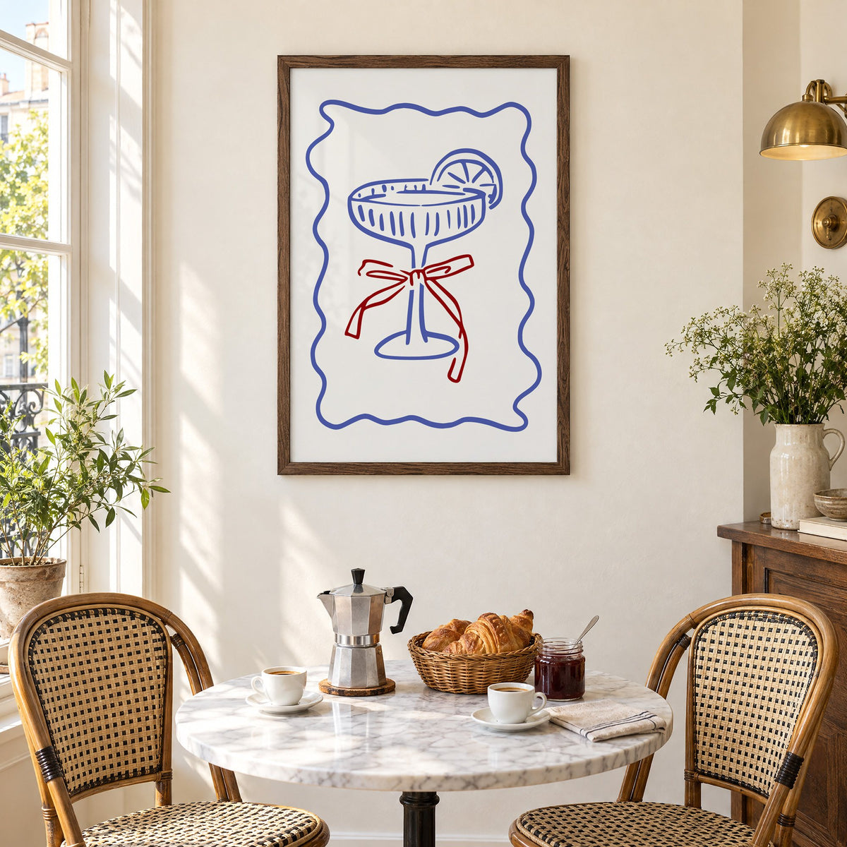 Blue coupe glass with red bow print in a walnut frame above a marble bistro table with croissants and rattan chairs