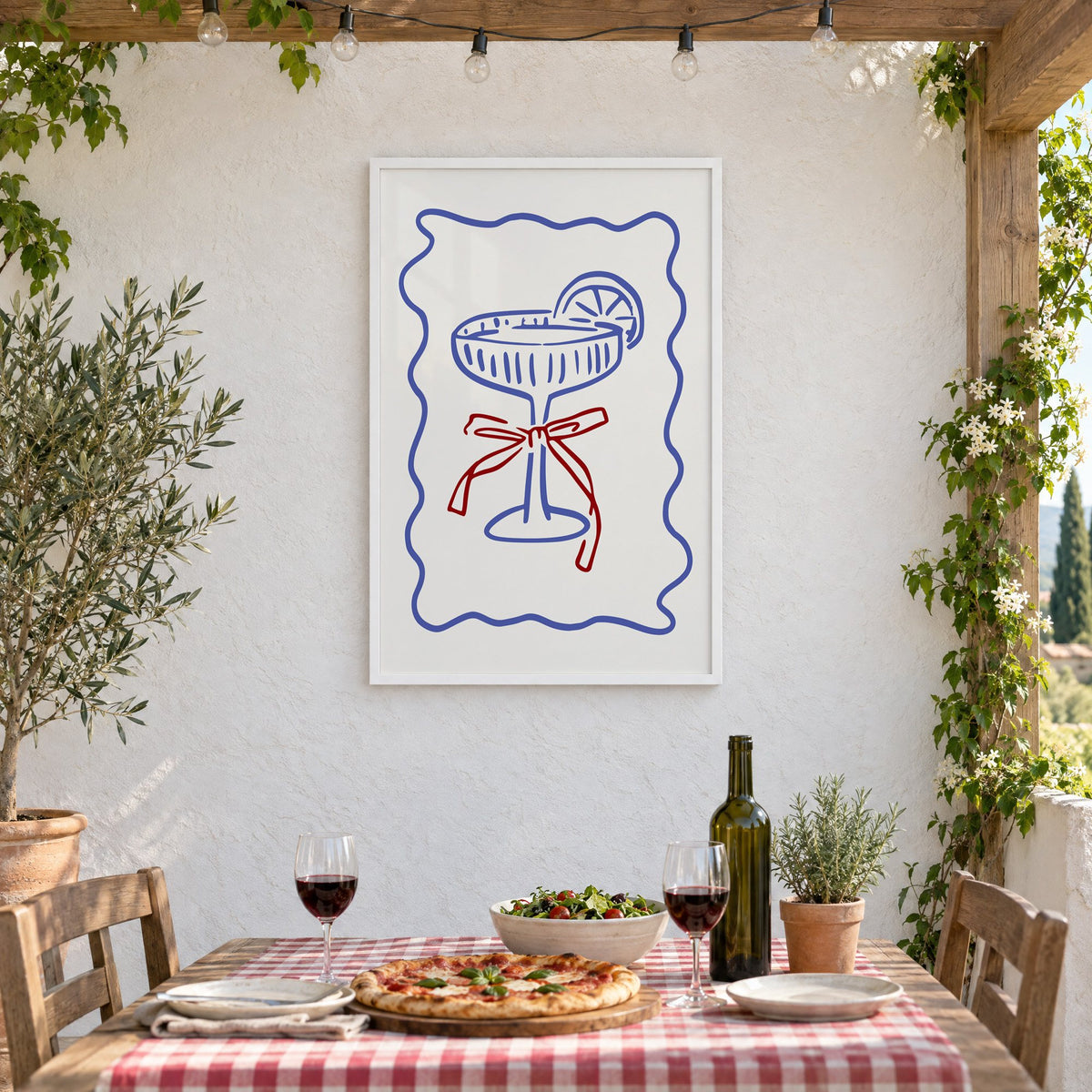 Blue coupe glass with red bow print in a white frame on a stucco wall above a checked-cloth table with pizza and red wine