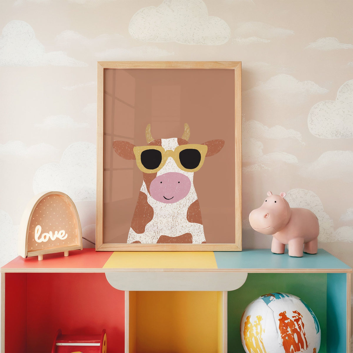 Cow in yellow sunglasses nursery print in an oak frame on a rainbow toy shelf against cloud wallpaper