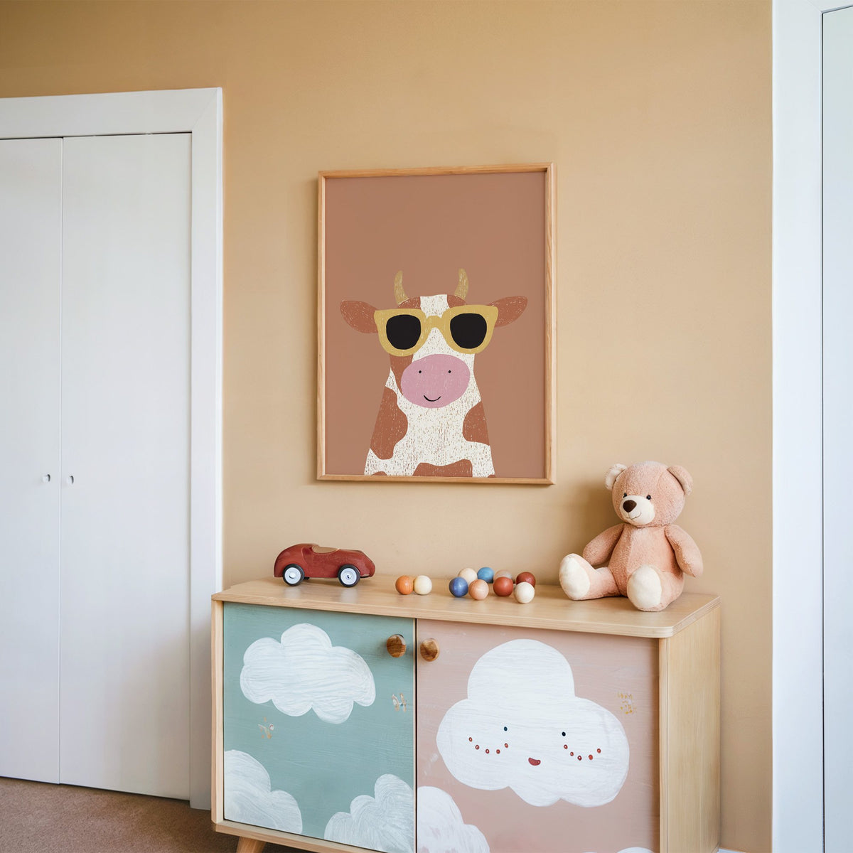 Cow in yellow sunglasses nursery print in an oak frame on a peach wall above a cloud-painted cabinet