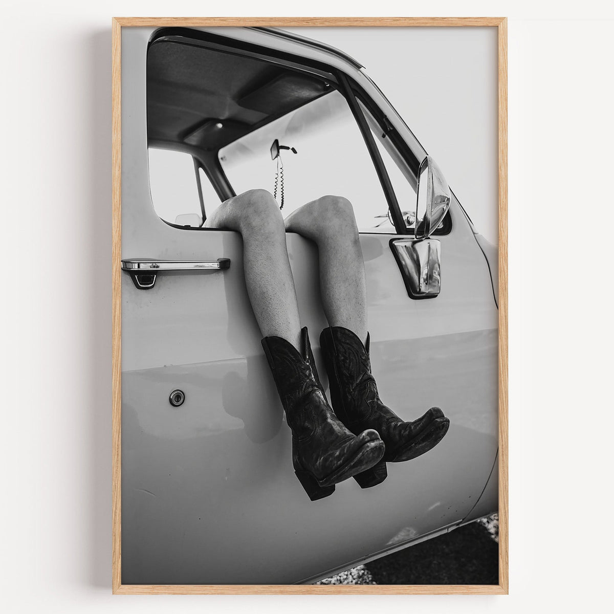 Cowboy boots dangling from a truck window print in a light oak frame on a plain white wall