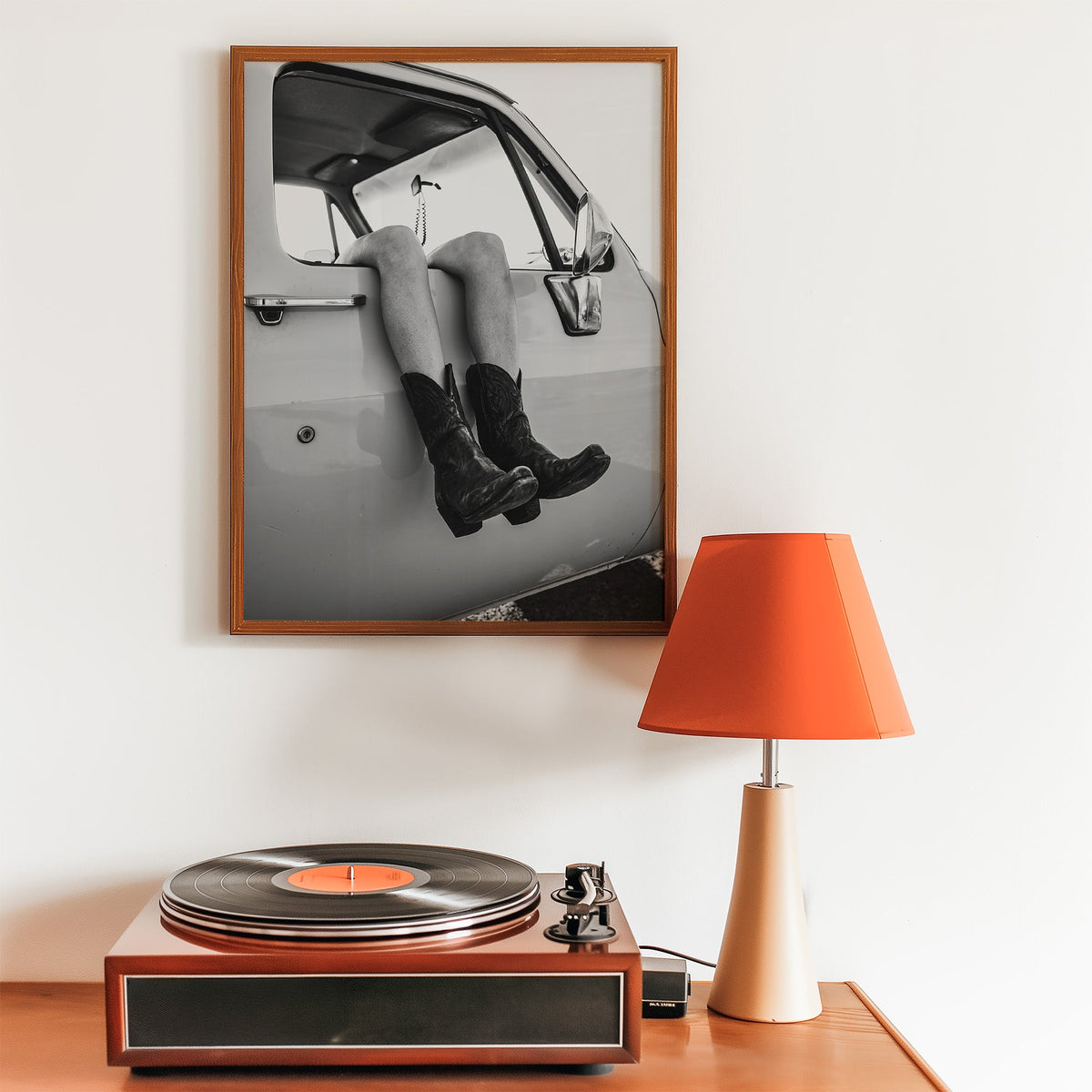 Cowboy boots dangling from a truck window print in a walnut frame above a record player and orange lamp