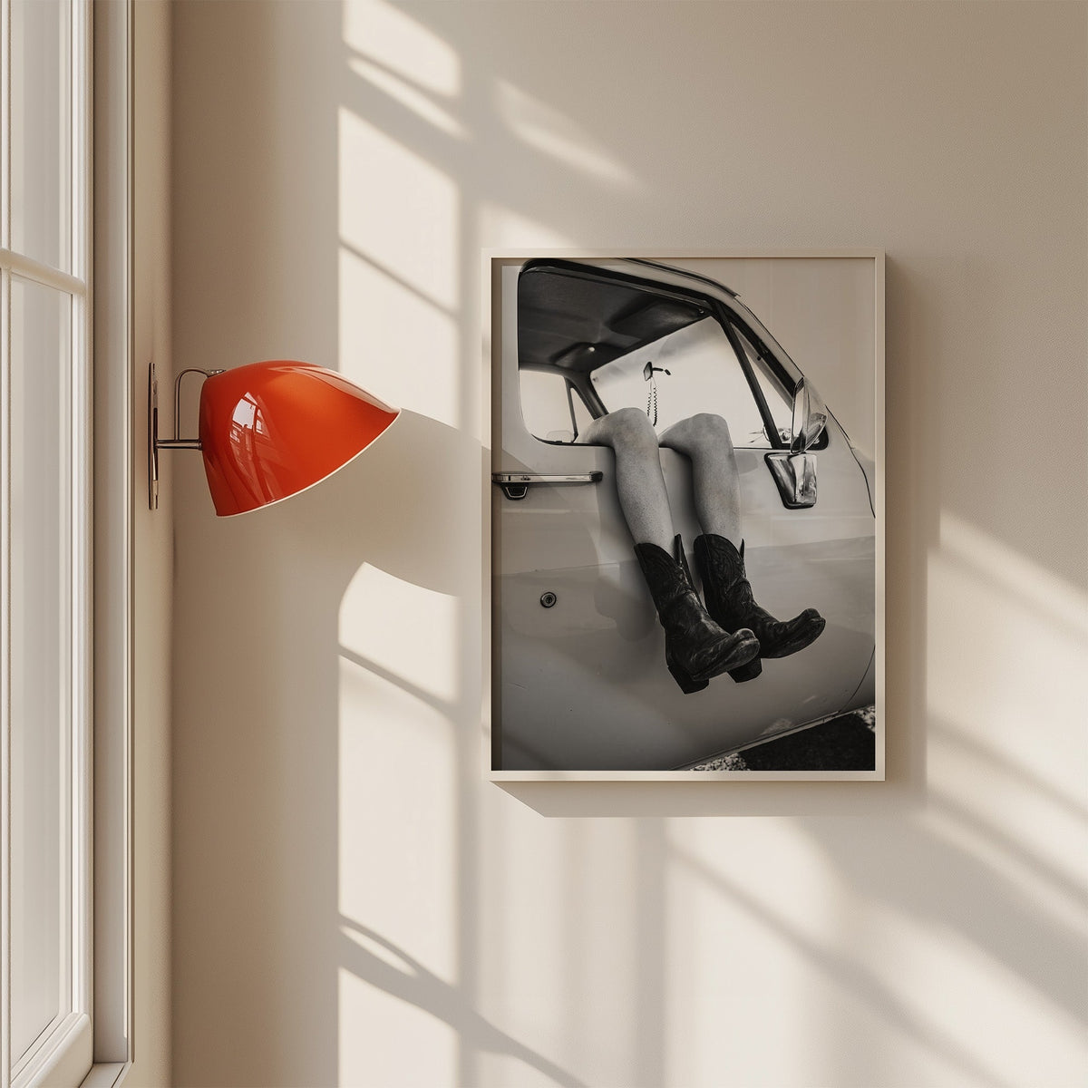 Cowboy boots dangling from a truck window print in a white frame on a sunlit wall beside a red sconce