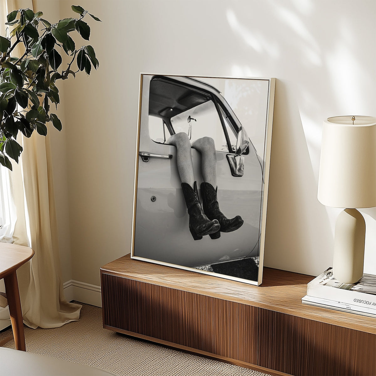 Cowboy boots dangling from a truck window print in a white frame on a walnut sideboard beside a ceramic lamp